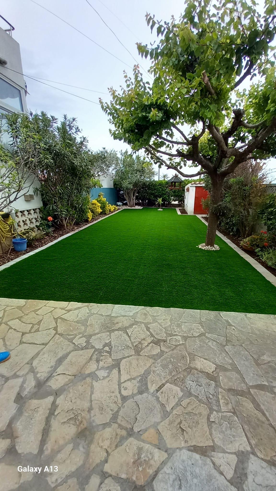 El mismo jardín terminado, con césped artificial uniforme entre los parterres y el pavimento de piedra